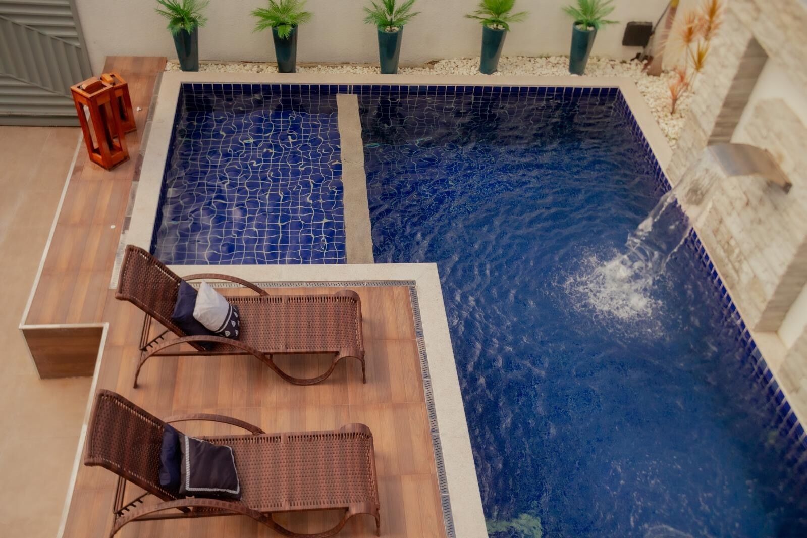 Une piscine rectangulaire aux carreaux de mosaïque bleus, entourée d’une terrasse en bois et agrémentée de deux fauteuils de jardin en osier.