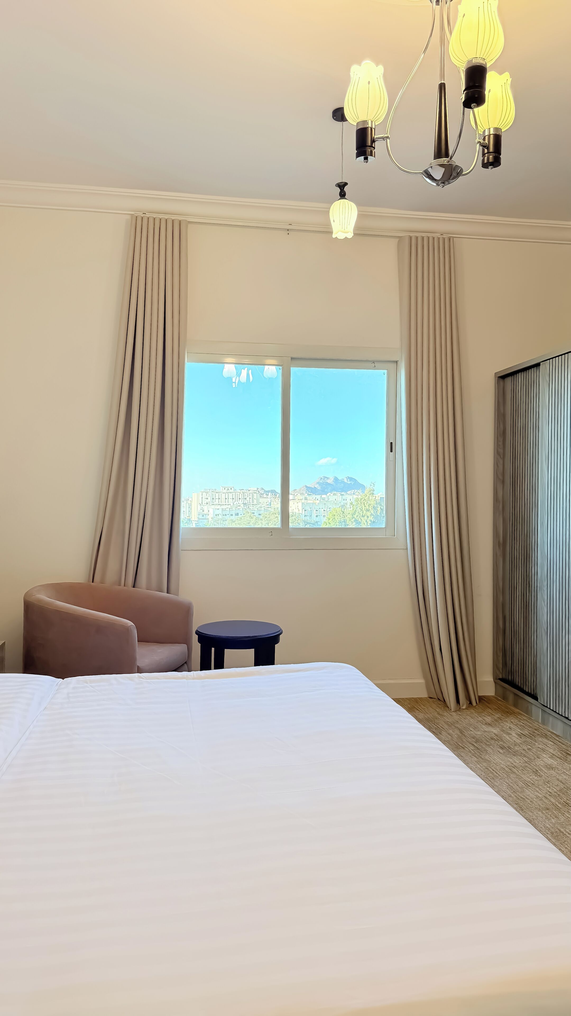 Habitación de hotel con una cama grande, una silla, una mesita y vista a un paisaje urbano a través de la ventana.