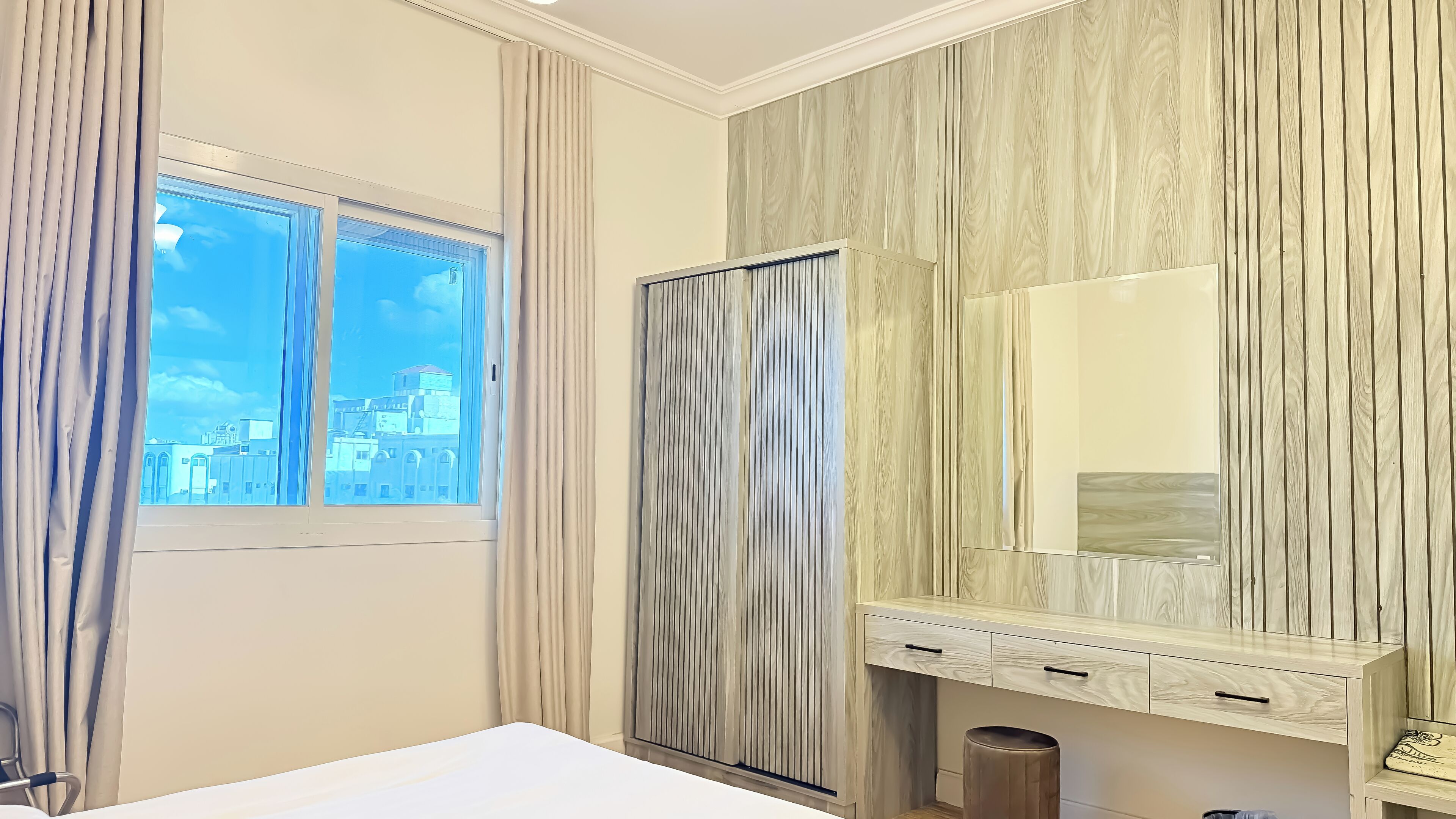 Una habitación de hotel con un ventanal grande, un armario, un espejo y una mesita de noche.