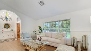 A spacious living room with a white sofa, a glass coffee table, and a large window with a view of greenery. - The Gardens Estate - Luxury Resort Retreat Near PGA Golf, Beaches, & Jupiter (West Palm Beach)