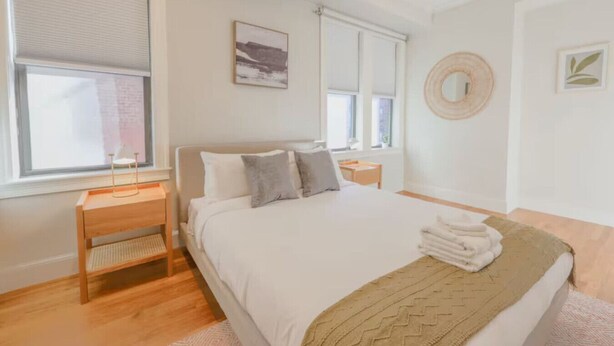 A bedroom with a bed, bedside tables, a window with blinds, a wall-mounted mirror, and a framed picture.
