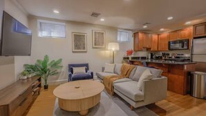 A modern living room with a sofa, armchair, and coffee table, a kitchen with wooden cabinets and appliances, and a flat-screen TV mounted on the wall.