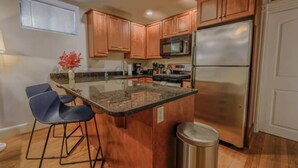 A kitchen with wooden cabinets, a stainless steel refrigerator, a microwave, and a granite countertop. There are two bar stools and a trash can.