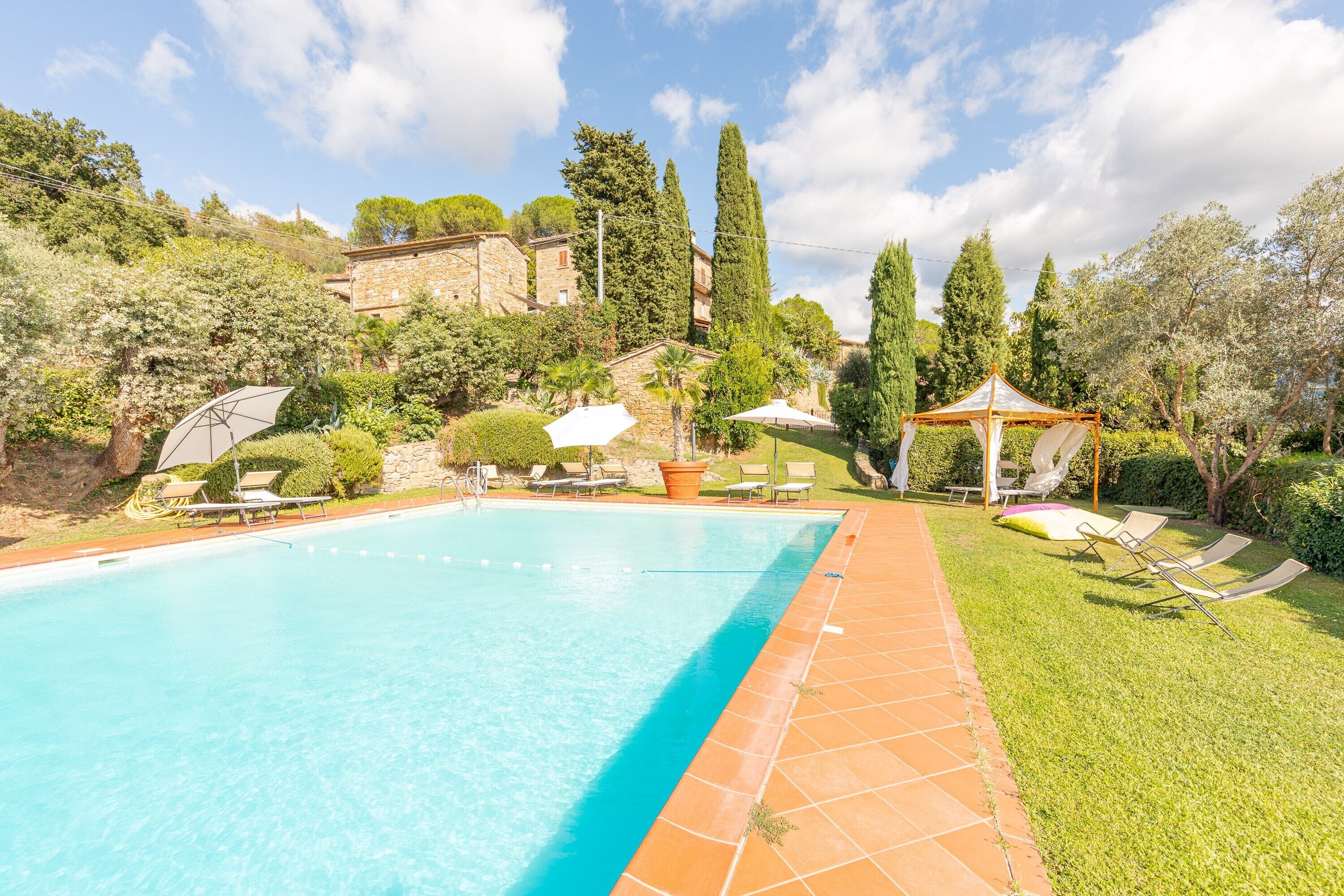 A swimming pool with lounge chairs and umbrellas, surrounded by greenery and stone buildings.