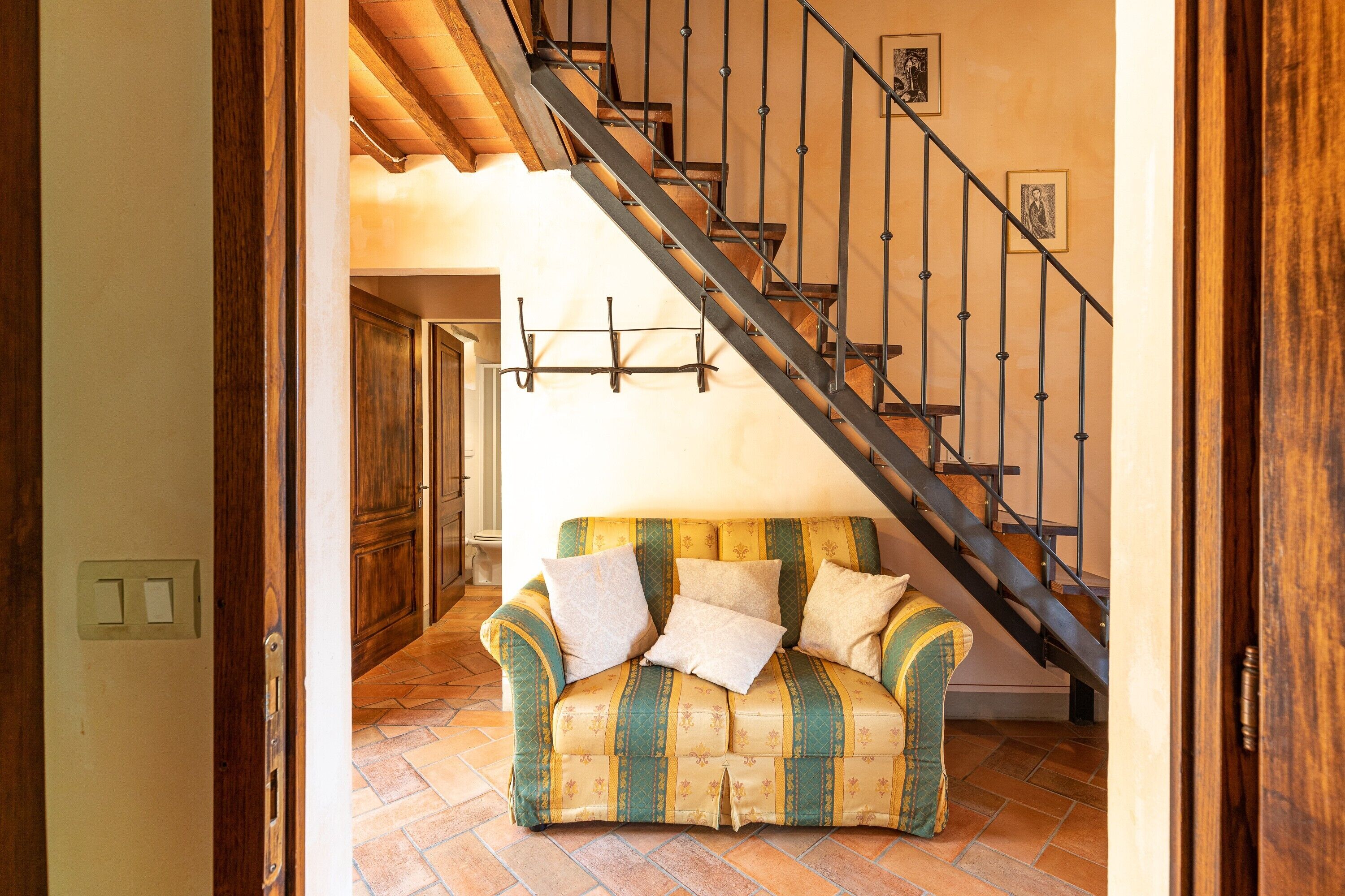 Un escalier avec une rampe en métal, une porte en bois et un canapé à motifs dans une pièce dont le sol est recouvert de carreaux.