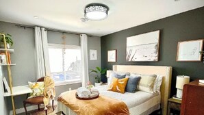 A bedroom with a large bed, a bedside table, a chair, a desk, and a window with curtains.