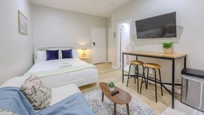 A modern hotel room with a bed, a small sitting area, a desk with stools, a TV, and a trash bin. - Old Charm Studio Apt near Back Bay (Boston)