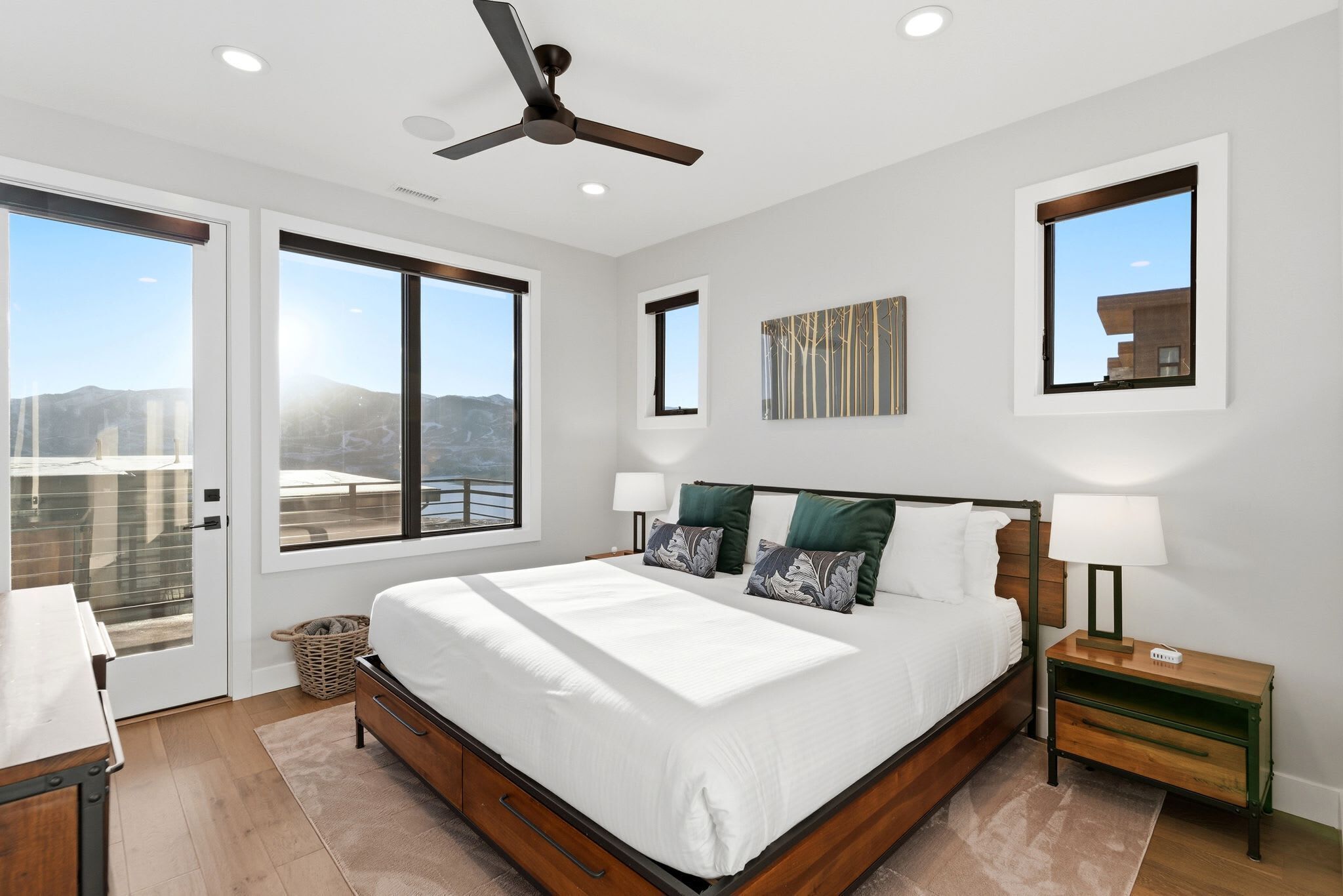 A modern bedroom with a large bed, two bedside tables, a ceiling fan, and large windows offering a view of the outdoors.