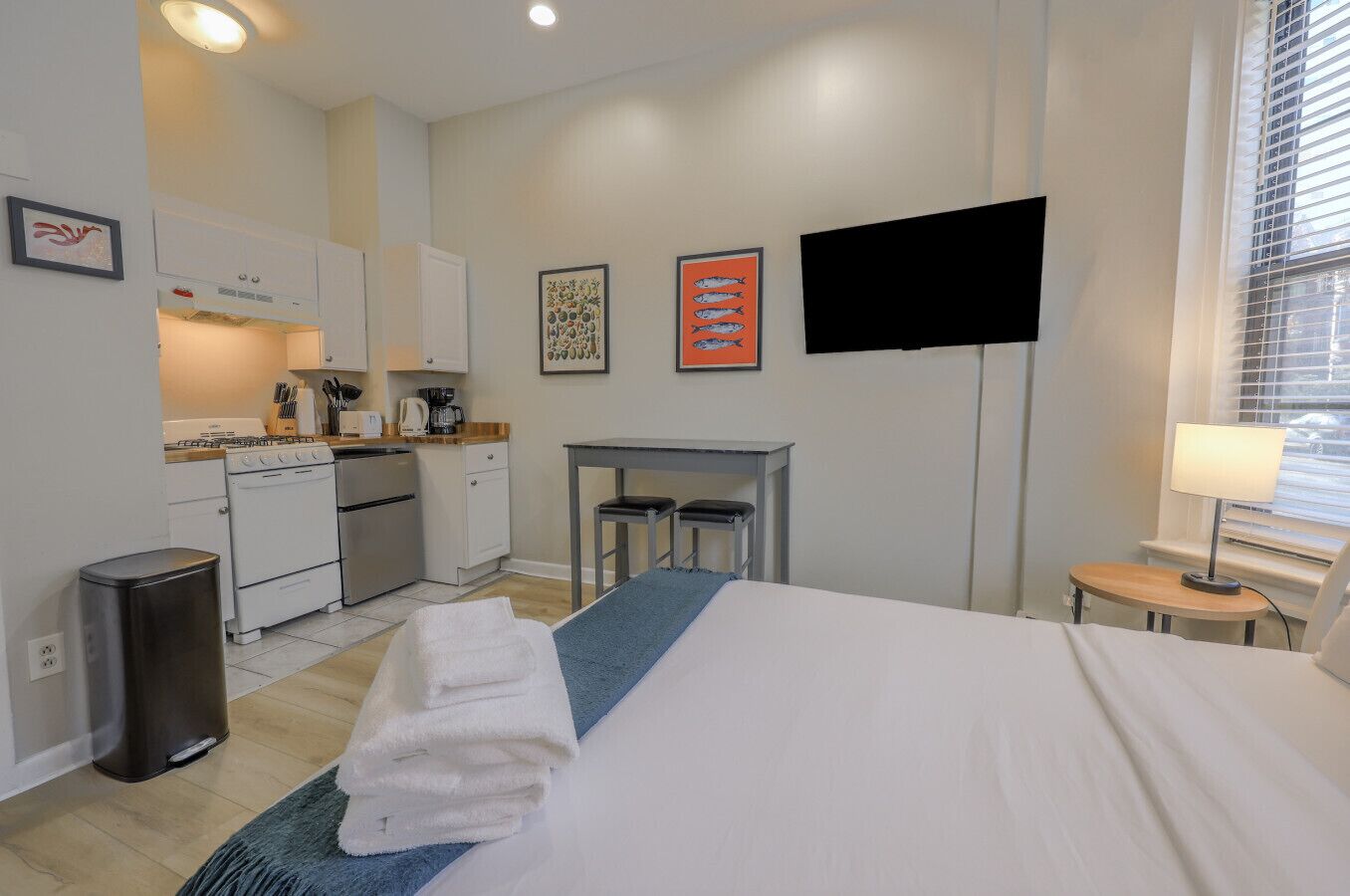 A compact hotel room with a bed, a small desk, a chair, a TV, and a kitchenette.
