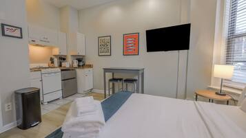 A compact hotel room with a bed, a small desk, a chair, a TV, and a kitchenette.