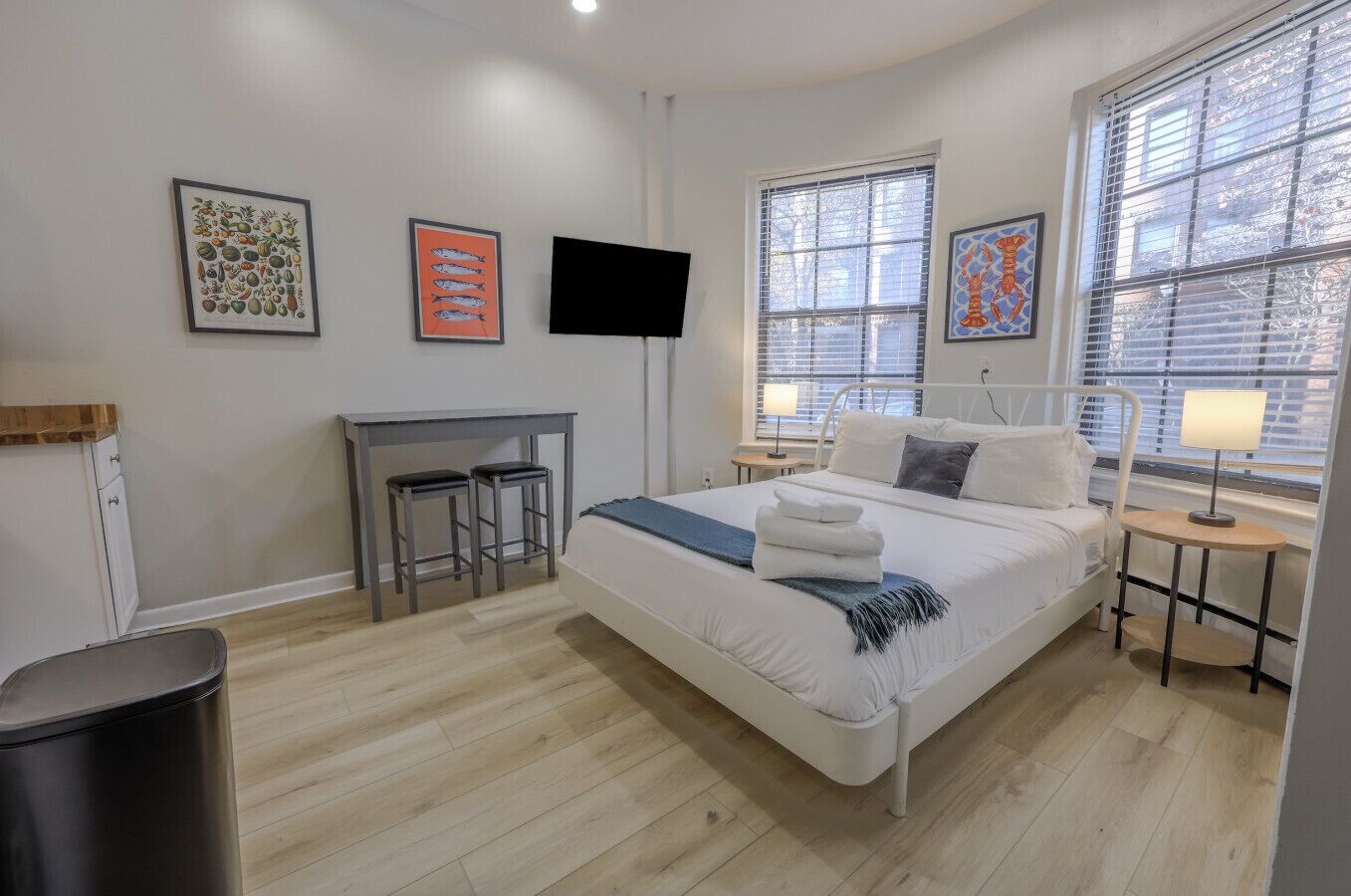 A modern hotel room with a bed, a desk with two stools, a TV, and artwork on the walls.