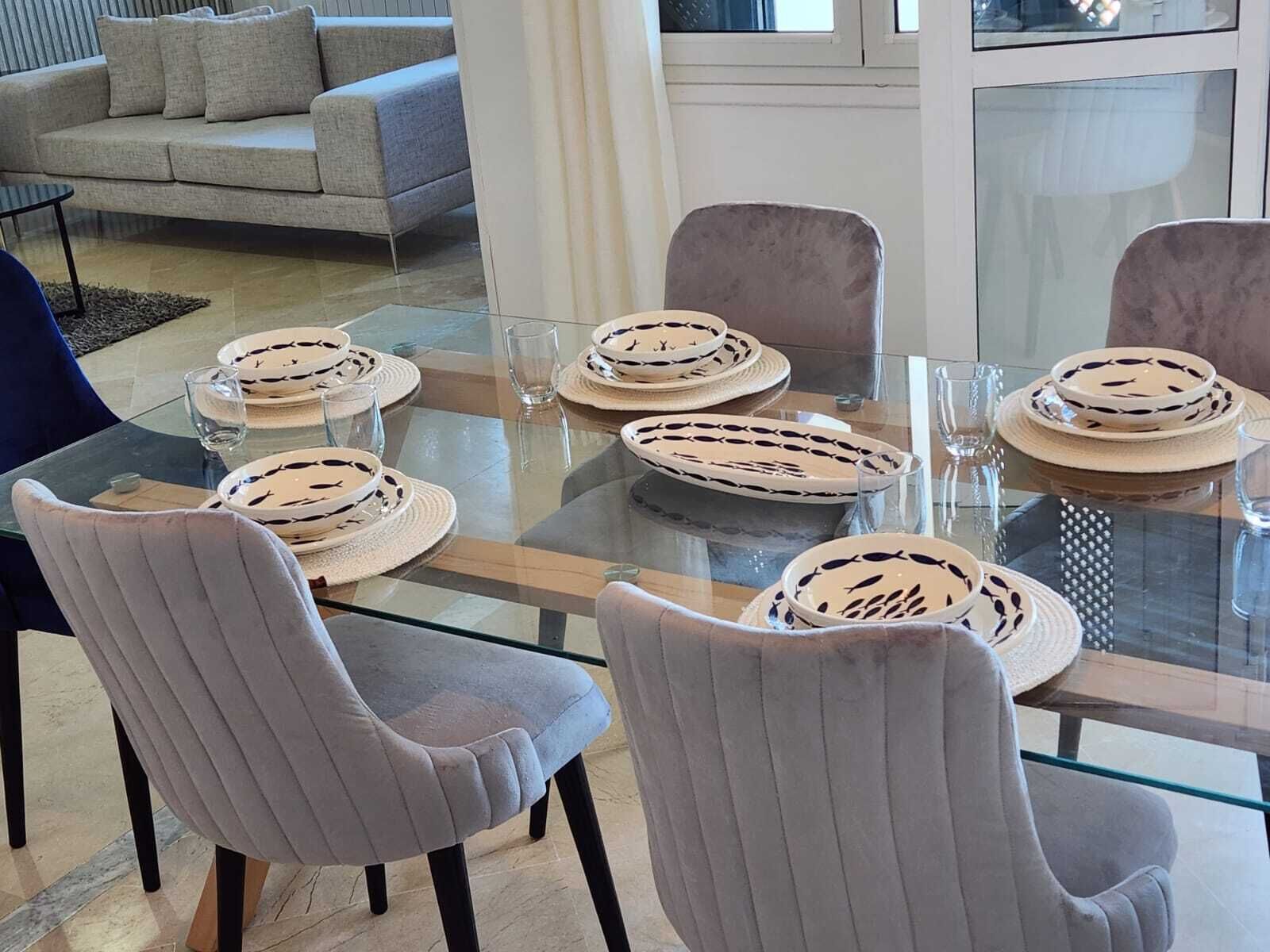 A dining area with a glass table, four grey upholstered chairs, and a grey sofa in the background.