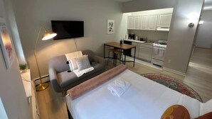 Et moderne hotelværelse med seng, skrivebord, sofa, fjernsyn og et tekøkken i baggrunden.