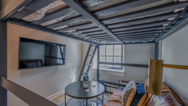 Ein kompakter Raum mit hohen Decken, einer Couch, einem kleinen Tisch, einem an der Wand befestigten Fernseher und einem Fenster mit Tageslicht.