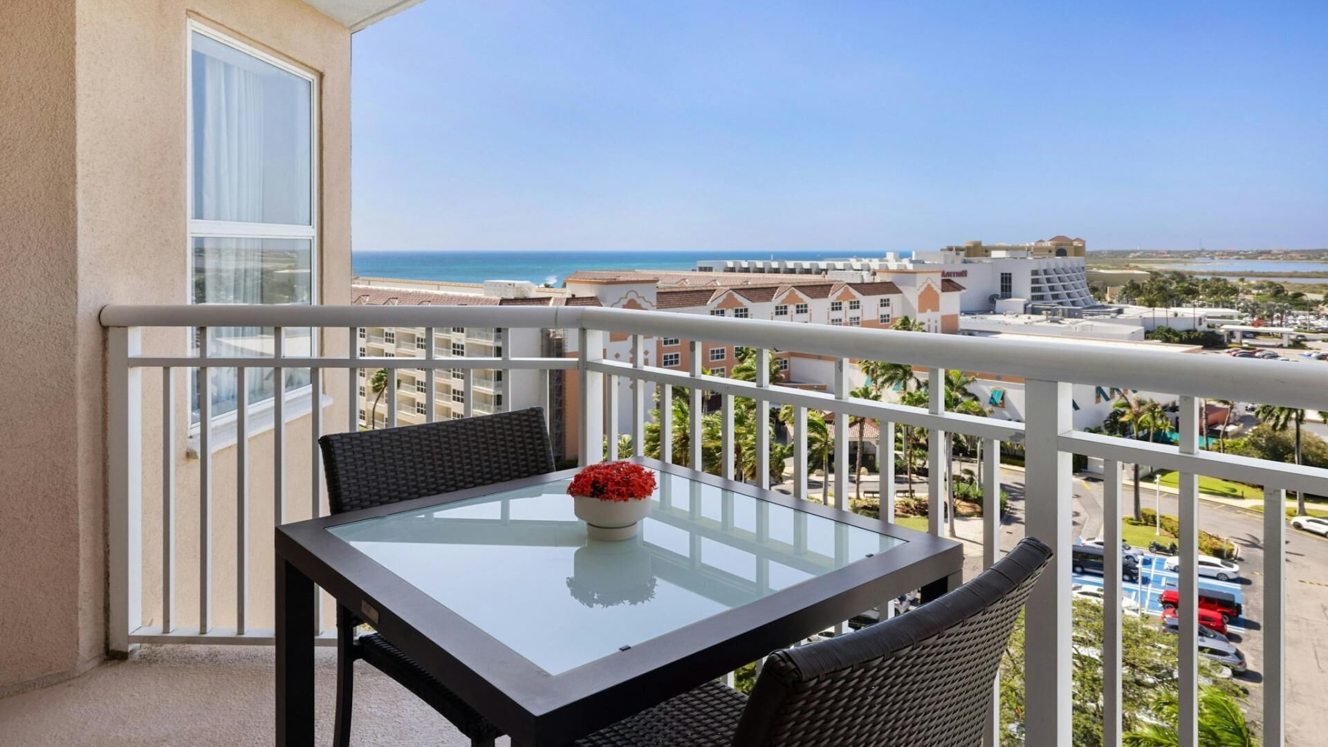 Un balcon avec une table et des chaises en verre, une plante en pot et une vue sur la plage et les bâtiments.