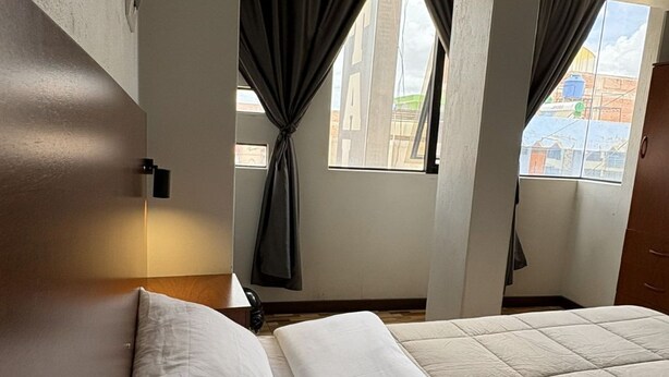 A hotel room with a bed, a nightstand, a lamp, and a view of buildings through the window. - Hostal - HS Merced (Juliaca)