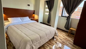 A hotel room with a large bed, wooden flooring, a nightstand, a small table, and a window with curtains. - Hostal - HS Merced (Juliaca)