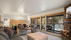 Een ruime woonkamer met een grote bank, gestreept fauteuils, een salontafel en een open haard. Er is een eethoek met een tafel en stoelen, en een grote schuifdeur die leidt naar een terras met buitenmeubels.