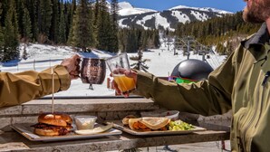 Eine Person stöstet mit einem Getränk bei einem Tisch an, auf dem ein Burger, ein Taco und ein Glas Orangensaft stehen, mit Blick auf einen verschneiten Berg und eine Seilbahn.