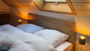 Une pièce chaleureuse avec un plafond en pente, une lucarne, un lit avec une literie blanche et une table de chevet avec une lampe.