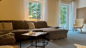 Un salon moderne avec un canapé d’angle, une petite table basse et un fauteuil. De grandes fenêtres sont ornées de rideaux, une lampe est posée sur une table d’appoint, et on peut admirer la verdure à l’extérieur.