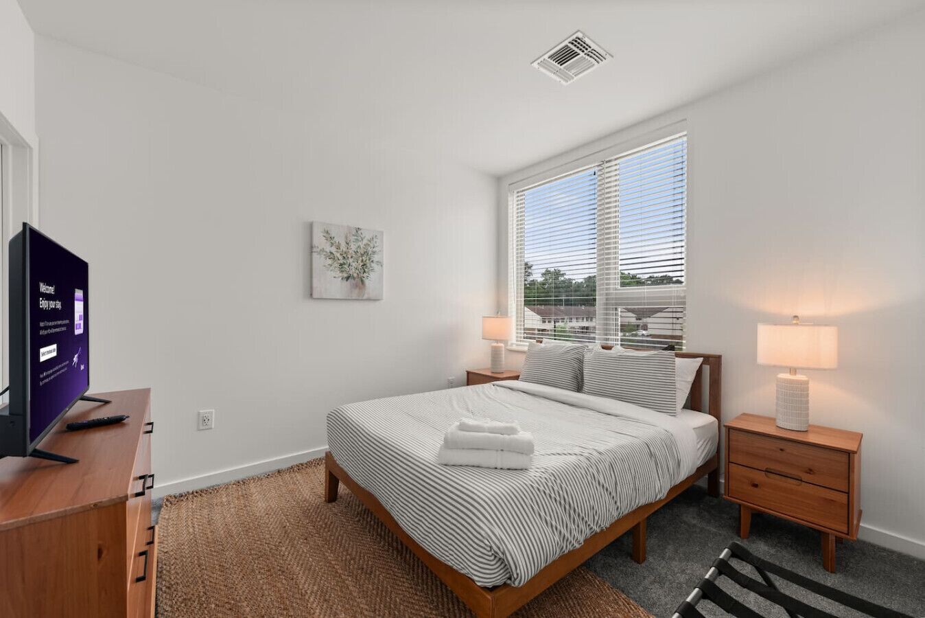 Un dormitorio moderno con cama, lámparas de noche, televisión y ventana con persianas.