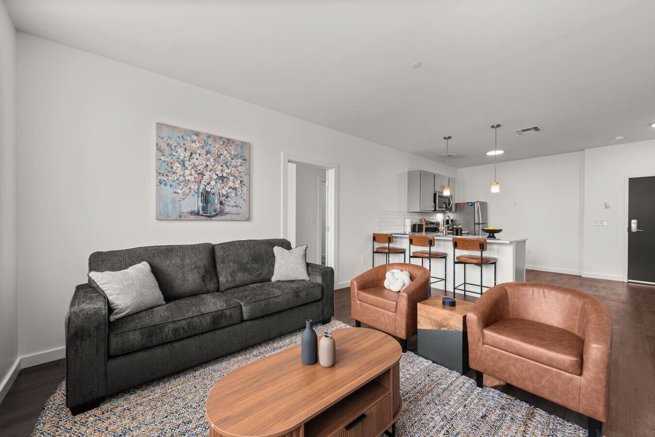 A modern living room with a grey sofa, a wooden coffee table, and two leather armchairs. A kitchen with bar stools is visible in the background.