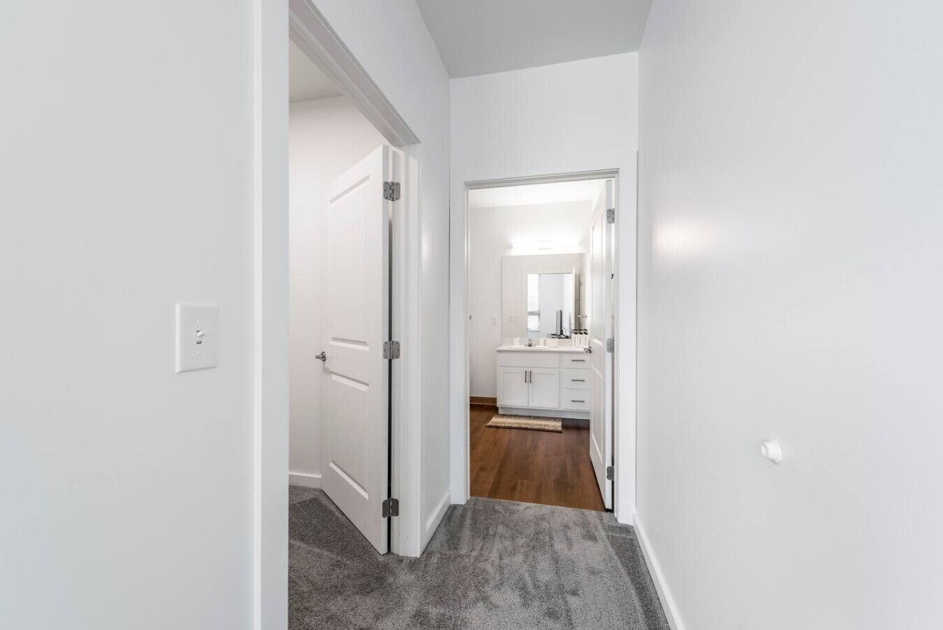 Un couloir menant à une salle de bain avec une porte blanche, un meuble-lavabo blanc et un miroir.