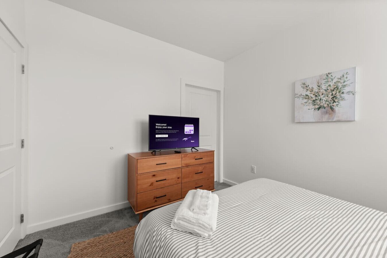 A hotel room with a bed, a wooden dresser, a television displaying a welcome message, and a painting on the wall.