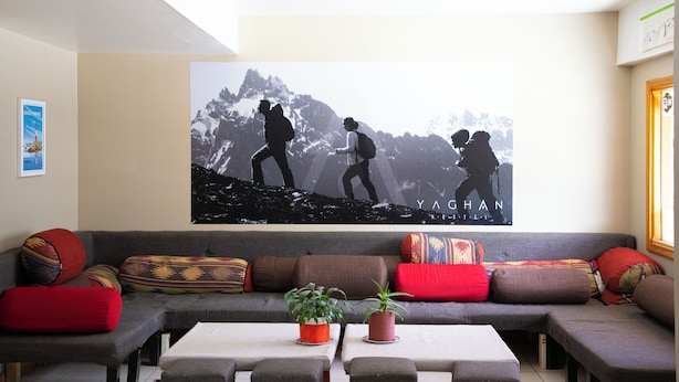 A living room with a large wall mural of hikers, a gray sofa with colorful cushions, and two potted plants on a coffee table. - YAGHAN HOSTEL (Ushuaia)