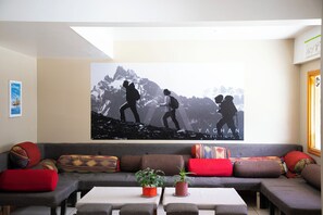 Un salon avec une grande fresque murale représentant des randonneurs, un canapé gris agrémenté de coussins colorés et deux plantes en pot posées sur une table basse.