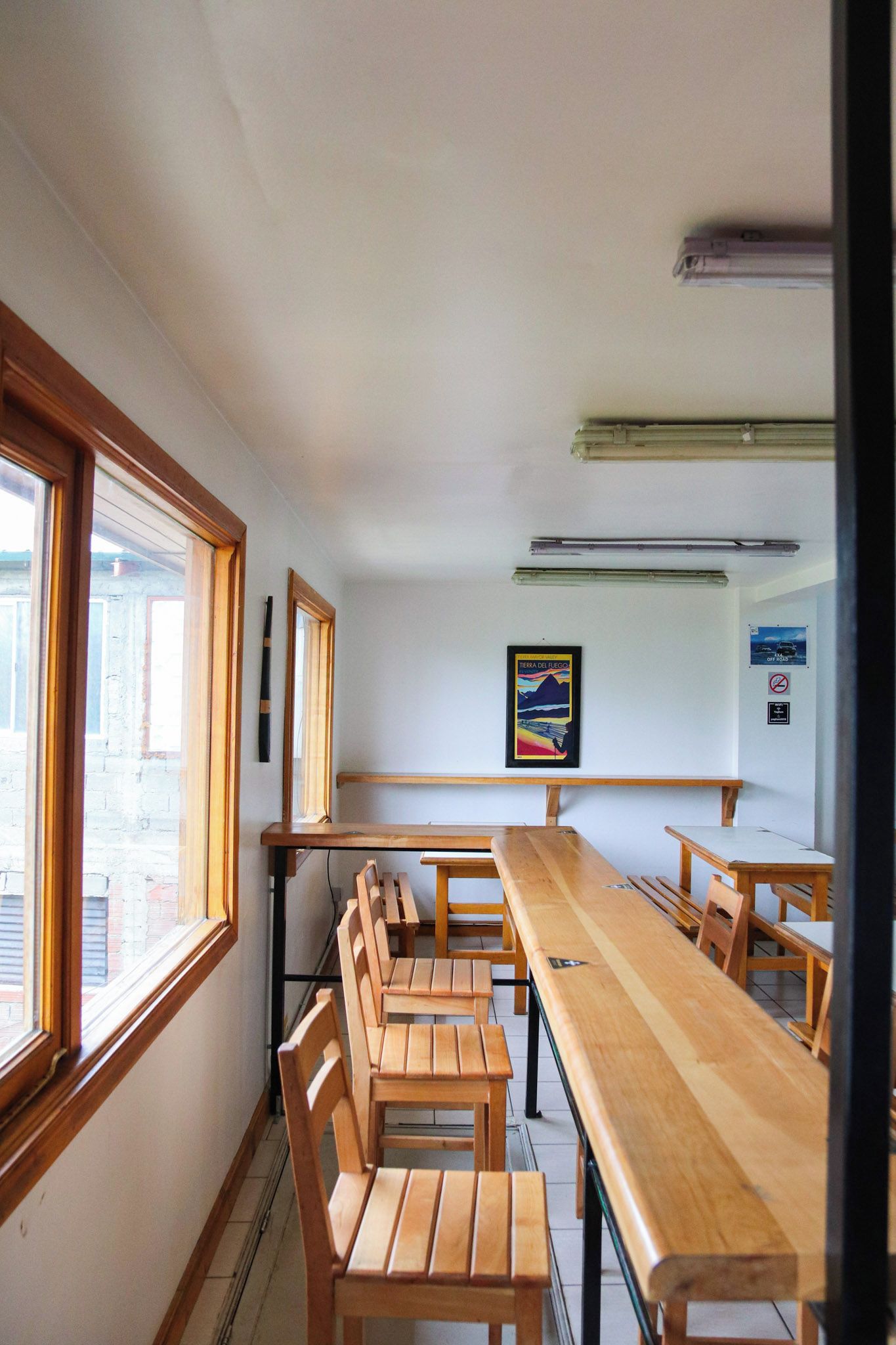 Un aula con pupitres y mesas de madera, un cuadro en la pared y una ventana a la izquierda.