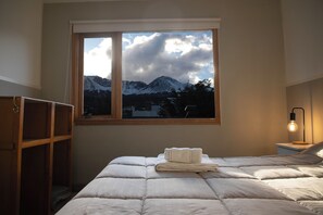 Un dormitorio con cama, ventana con vista a la montaña, mesita de noche con lámpara y armario de madera.
