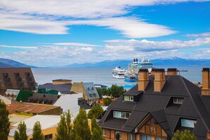 Eine Küstenstadt mit vor Anker liegendem Kreuzfahrtschiff, mit moderner und traditioneller Architektur, und freiem Blick auf Meer und Berge im Hintergrund.