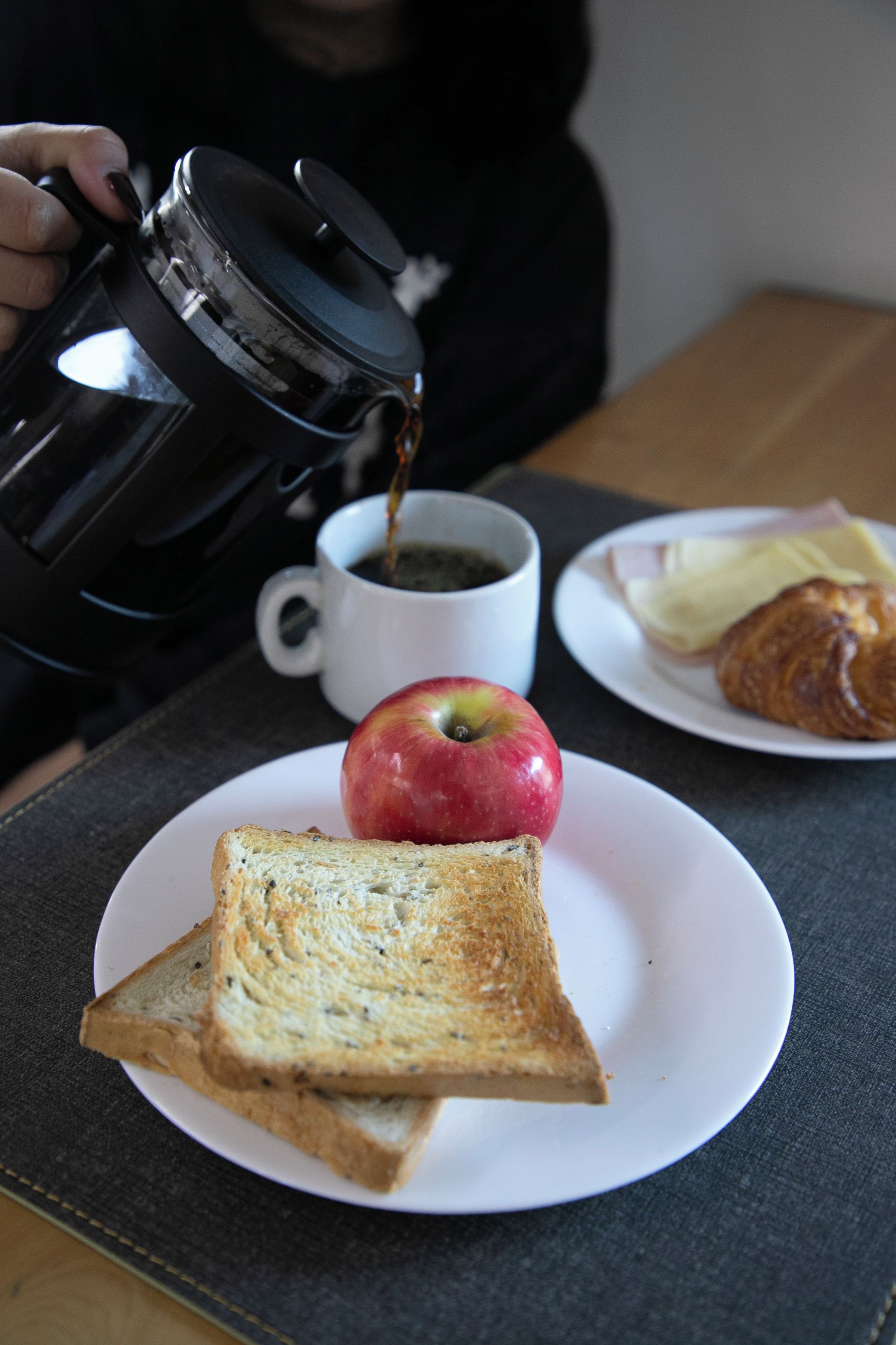 A table with a white plate containing toast, an apple, and a croissant, and a white mug with a beverage.