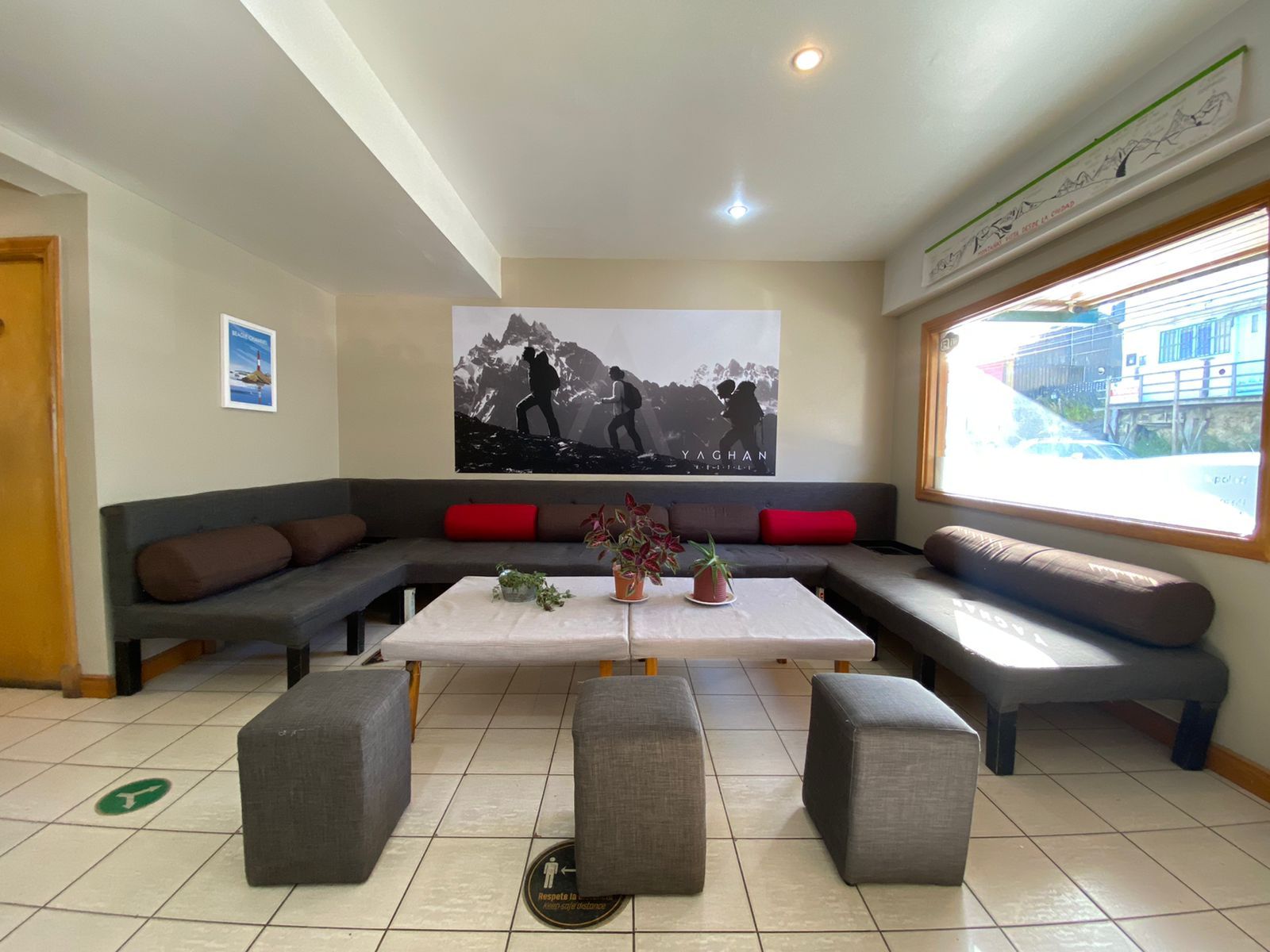 Un moderno espacio de descanso con un amplio sofá modular, una mesa de centro y dos otomanas. Hay un cartel en la pared y una ventana que ofrece vistas al exterior.