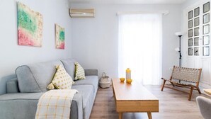 Ein modernes Wohnzimmer mit grauem Sofa, einem Holztisch und einem Korbstuhl. An der Wand befinden sich abstrakte Gemälde und ein Fenster mit weißen Vorhängen.