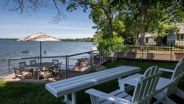 Un patio junto al lago con mobiliario exterior, una mesa de comedor y una sombrilla grande.