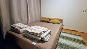 Ein ordentlich bezogenes Bett mit einer braunen Tagesdecke und Kissen, eine gefaltete Decke daneben und ein Streifenmuster-Teppich auf dem Boden.