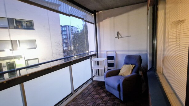 Ein Balkon mit blauem Stuhl, einem kleinen weißen Tisch und einem Fenster mit Rollläden.