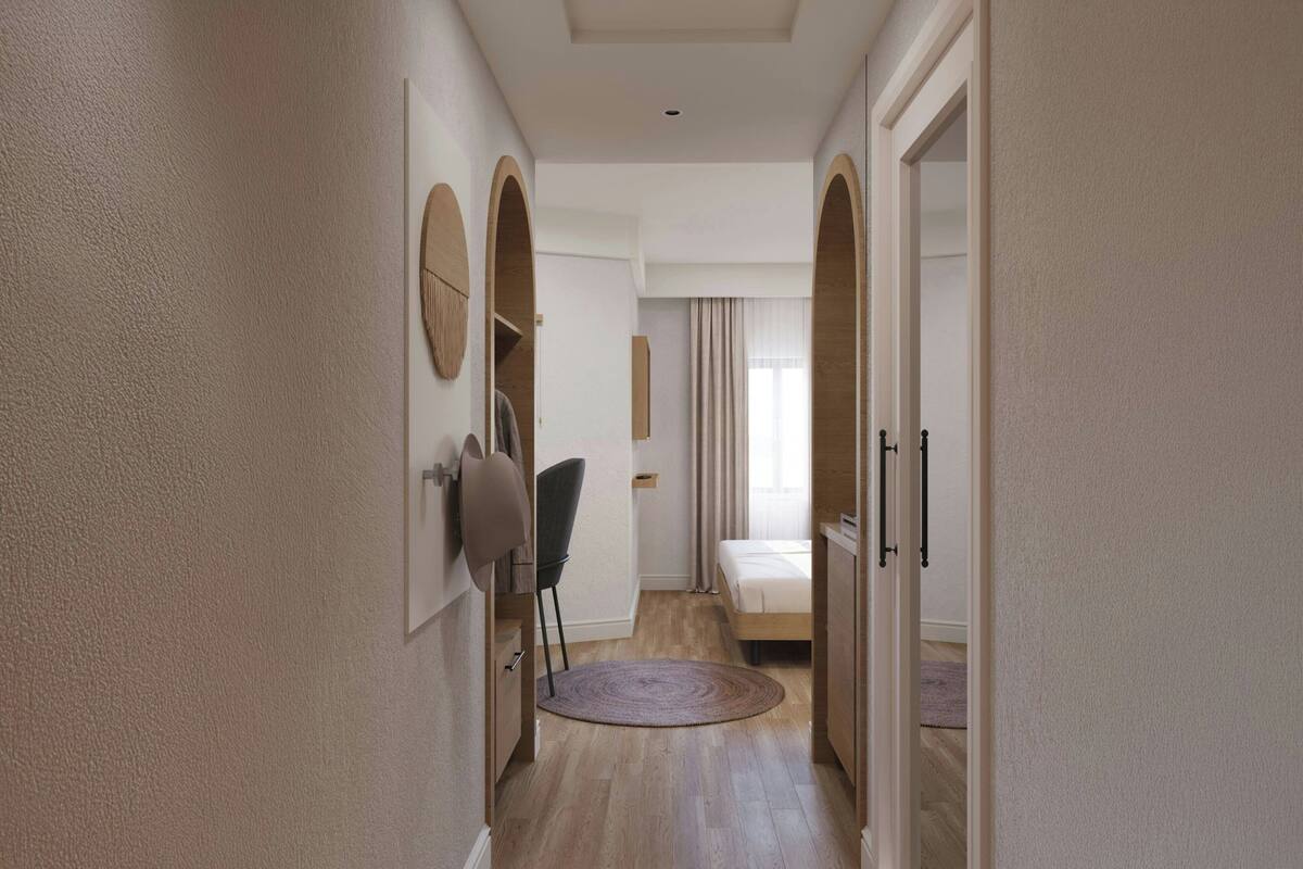 A narrow hallway leading to a bedroom with a bed, a chair, and a round rug.