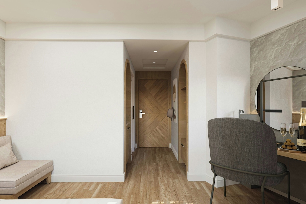 A modern hotel room with a wooden floor, a grey upholstered chair, a bed, a desk with a lamp, and a large mirror.