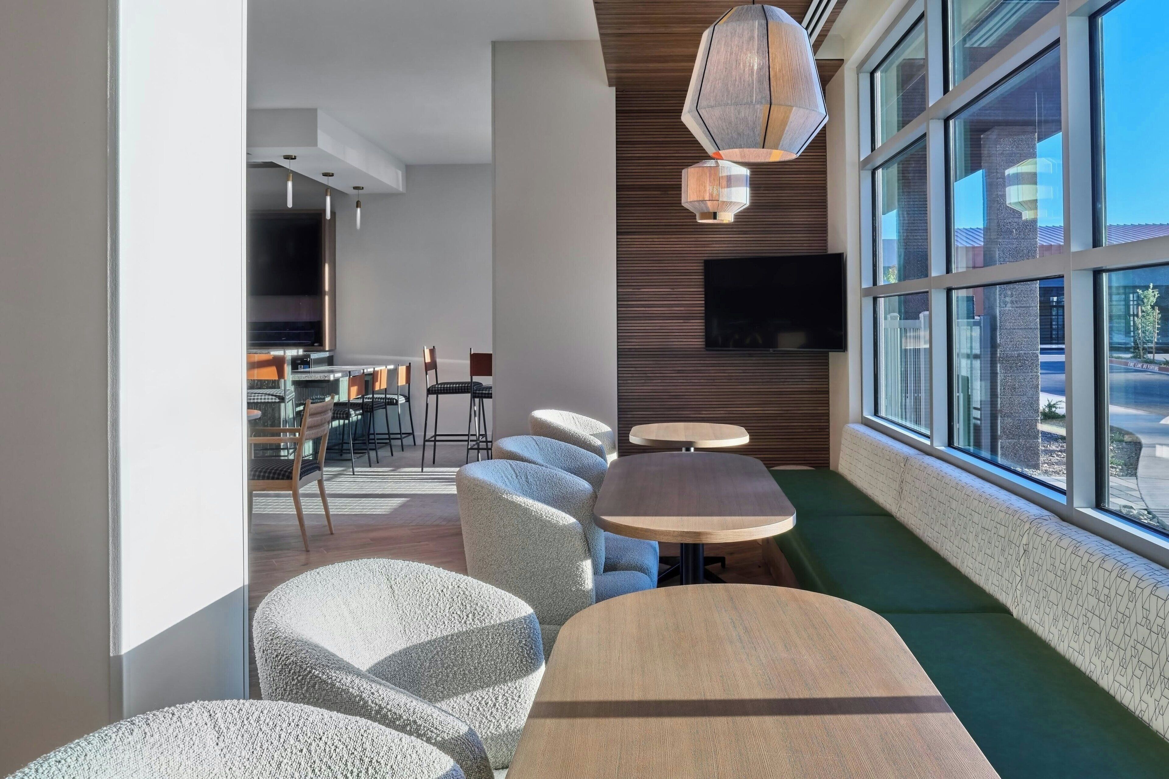 A modern interior with round tables and cushioned seating, a flat-screen TV mounted on the wall, and large windows offering a view outside.
