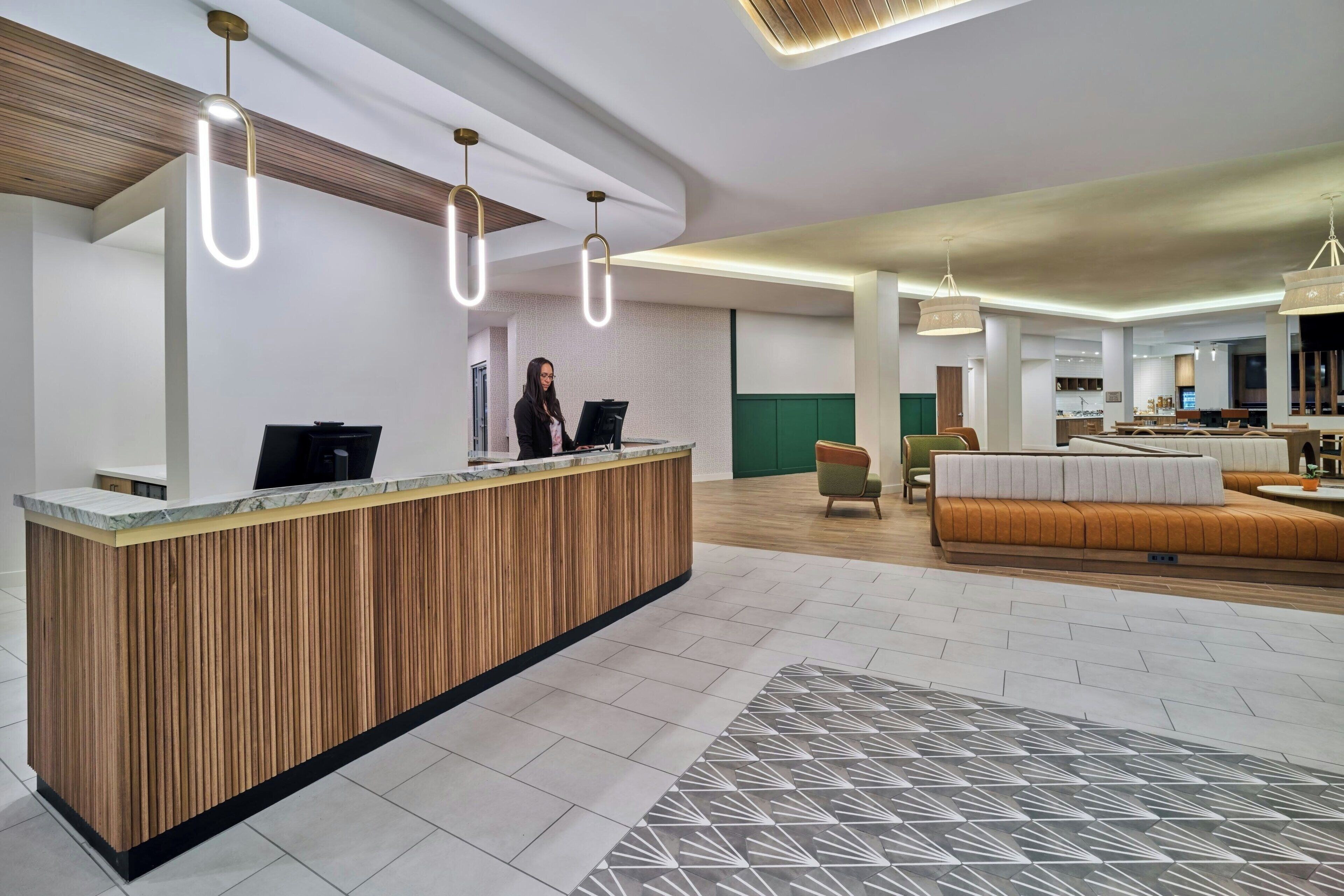 A reception desk with a wooden finish, a woman standing behind it, and a modern interior with a sofa and dining area in the background.
