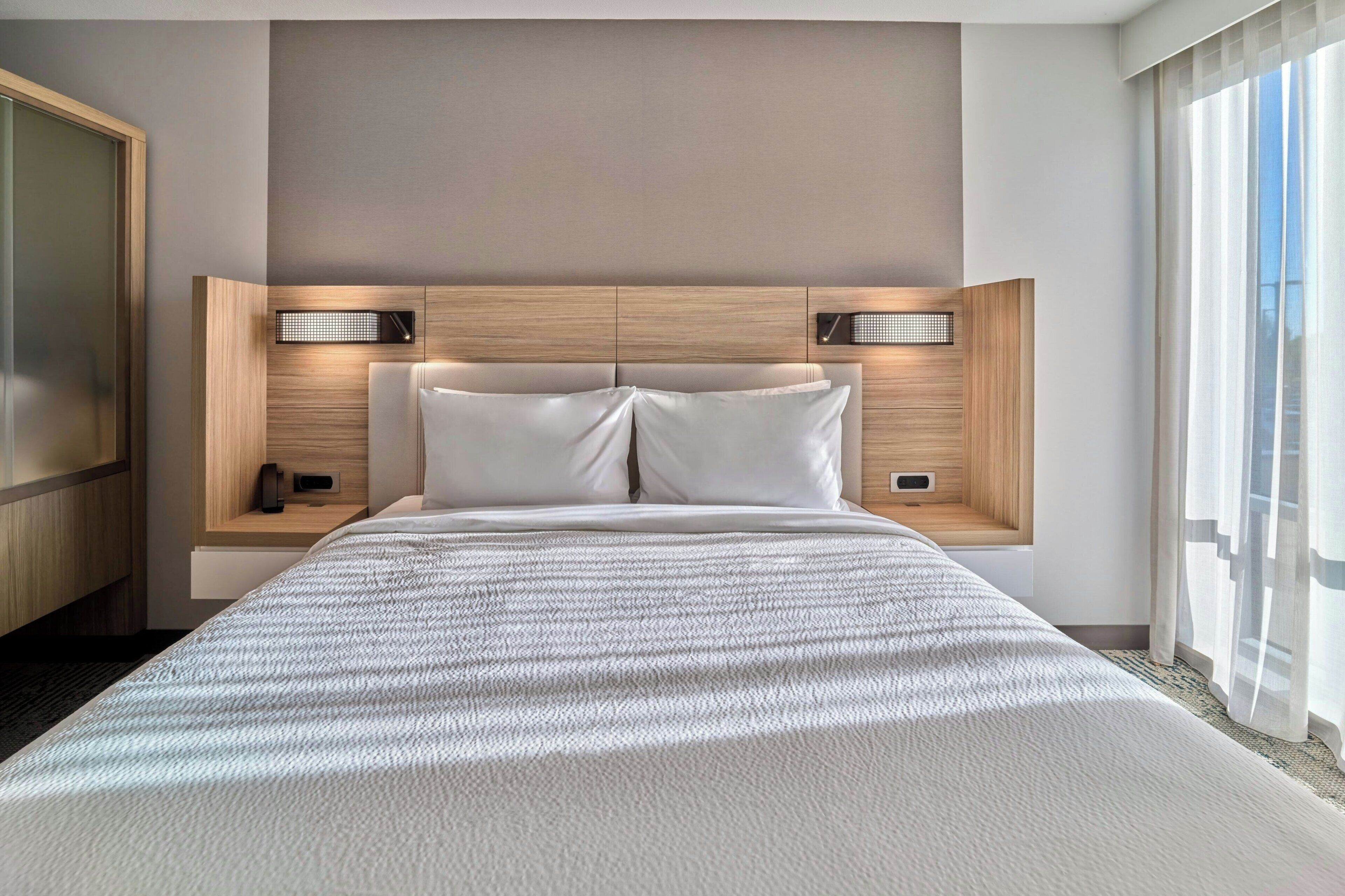 A modern hotel room with a large bed, two bedside tables, a built-in heater, and a large window with curtains.