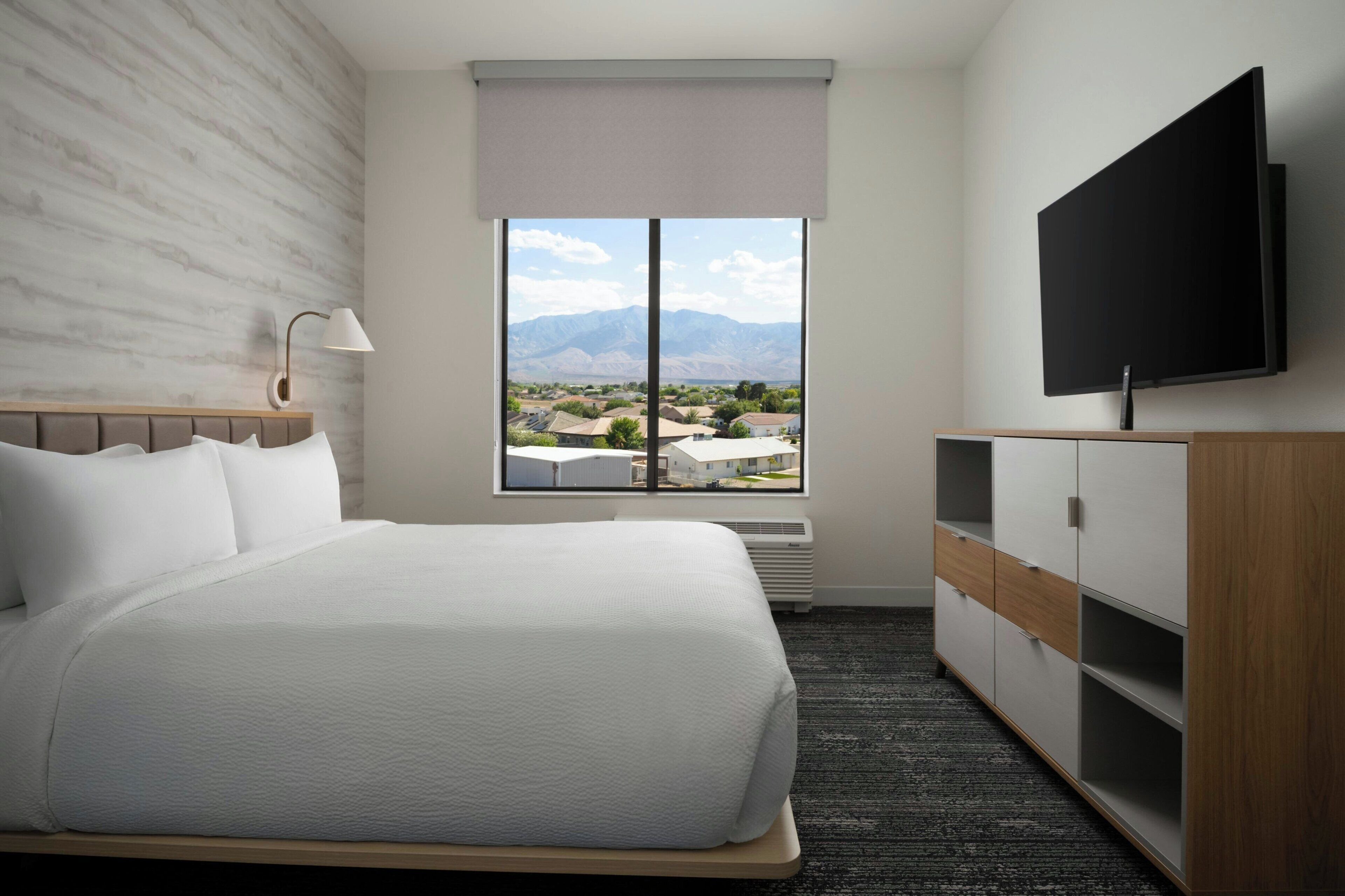 Una habitación de hotel con cama, televisión, ventana con vistas a la montaña y lámpara de pared.