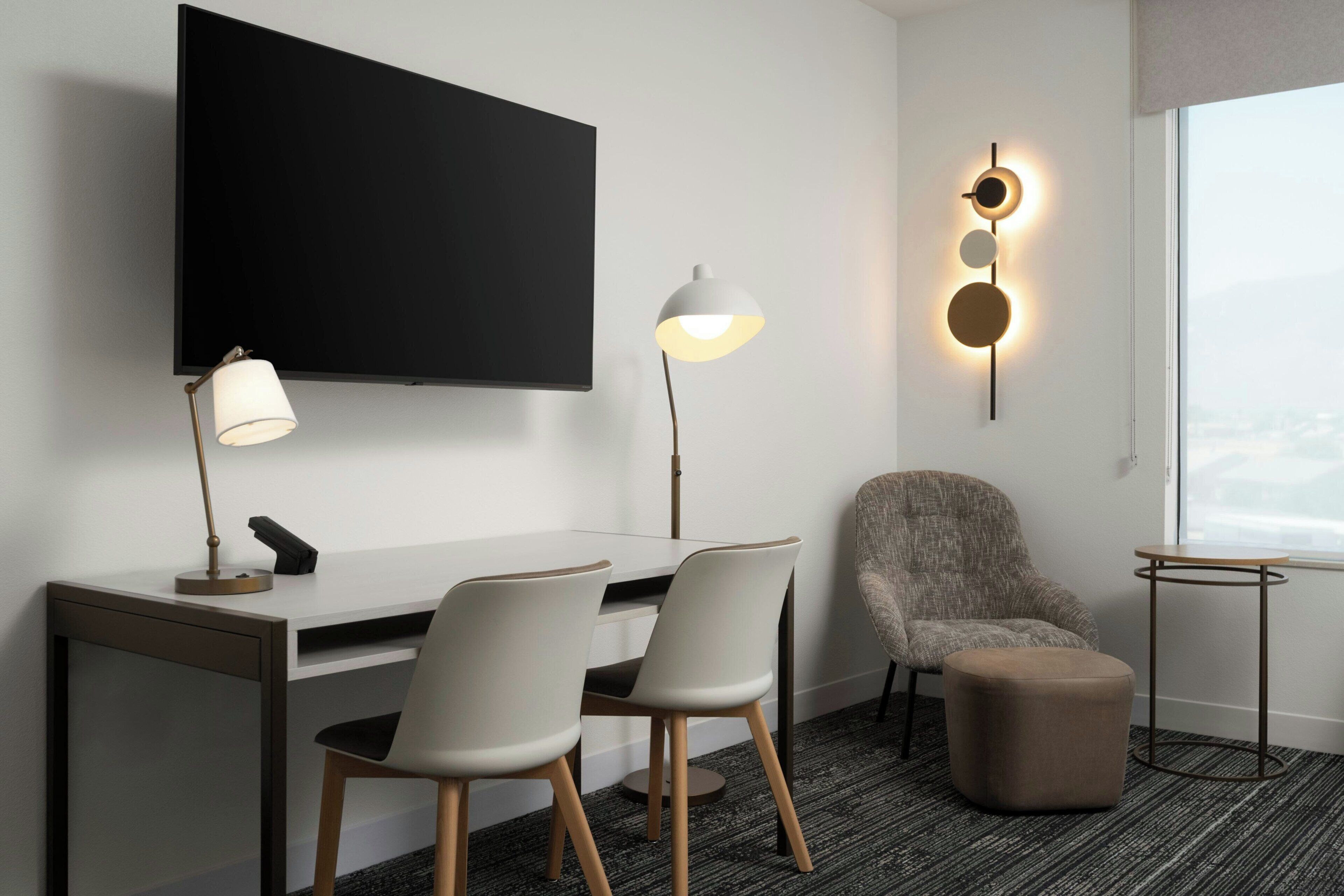 A modern hotel room with a large flat-screen TV, a desk with two chairs, a round ottoman, a side table, and a wall-mounted light fixture.