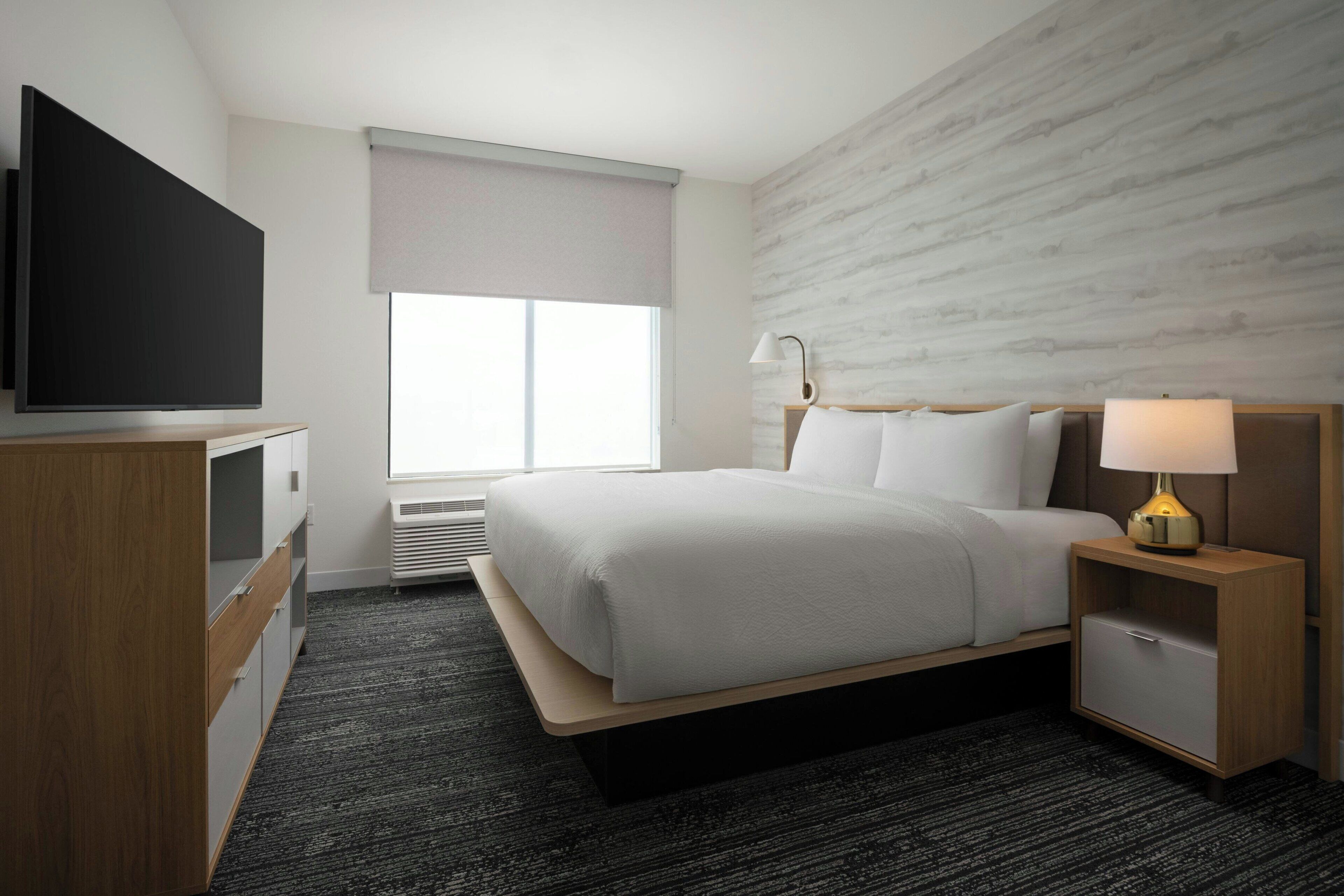 Une chambre d’hôtel comprenant un lit, une télévision, une table de chevet avec une lampe, une armoire et une fenêtre avec un store.