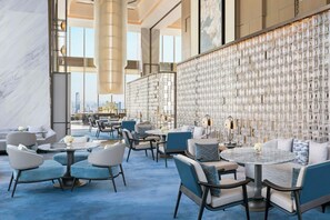 A modern hotel lobby with seating arrangements, a decorative wall, and a large window offering a city view.