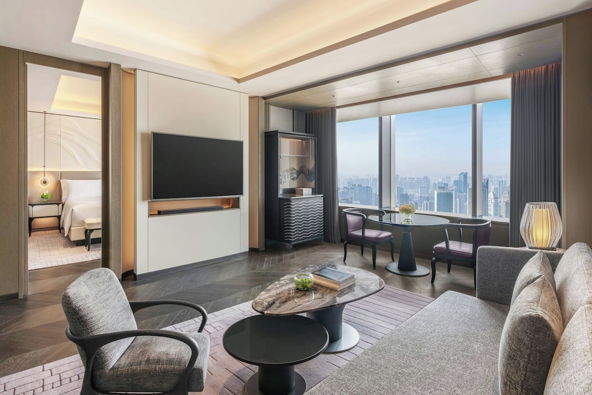 A modern hotel room with a large TV, a dining area, a sofa, and a view of the city.